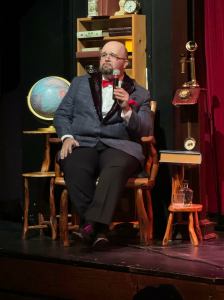 Live action of Kevin portraying the role of criminologist, sitting on a chair on stage in front of a bookcase and globe.